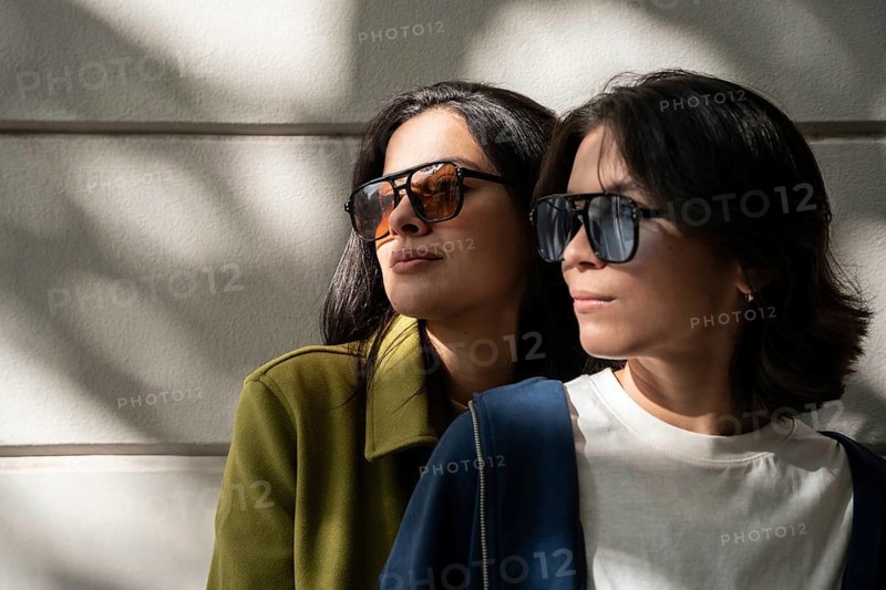 A fashionable lesbian couple wearing stylish sunglasses basks in warm sunlight, casting soft shadows on the wall. Their vibrant attire complements the serene, sunny setting