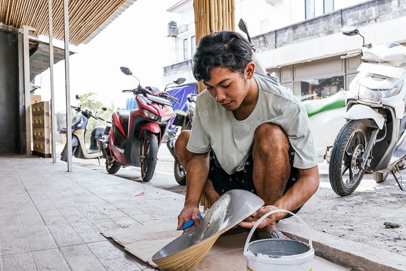 A Balinese artist skillfully paints a sculpture outdoors, blending traditional techniques with natural surroundings. Vibrant motorcycles are seen in the background, adding a modern touch