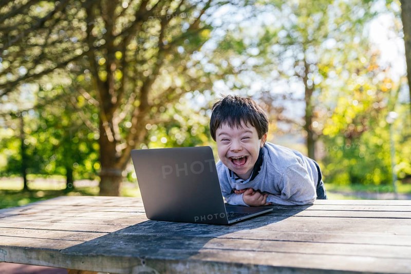 A cheerful child with Down syndrome is smiling joyfully at a laptop while sitting at a wooden table in a sunlit park. The image conveys happiness and technology