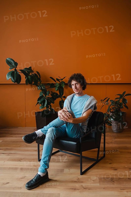 A young man with curly hair sits in a stylish chair in a contemporary room. The space features a warm, orange wall and lush green plants, creating a relaxing atmosphere