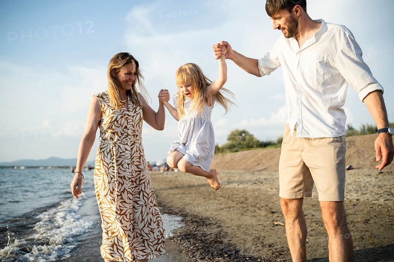 A cheerful family enjoys their time by the sea in Italy, as the parents swing their daughter into the air while walking along the shoreline. The child laughs with excitement and joy