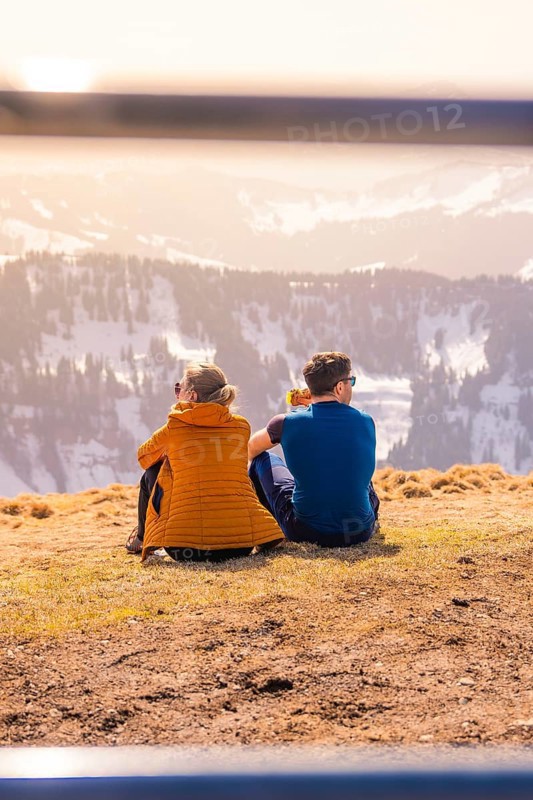 Couple sitting on a mountain peak with a view of the sunny winter valley, Hochgrat Mountains, Allgäu Alps, Oberreute, Germany