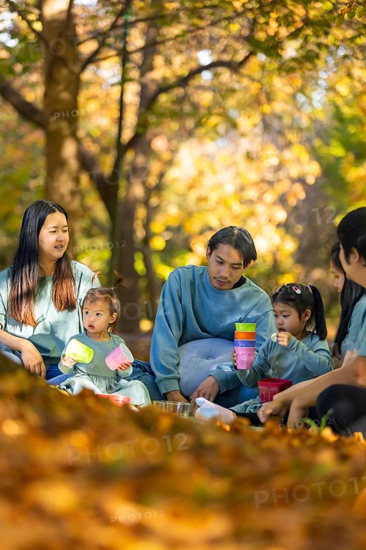 An Asian family enjoying a peaceful picnic surrounded by autumn foliage. They are sitting on a blanket in a park, sharing food and spending quality time together in the crisp fall air