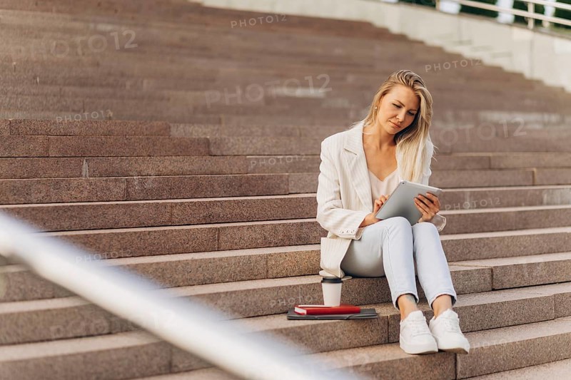 A woman in a white blazer and jeans sits on outdoor steps, using a tablet. Beside her are a takeaway coffee cup and a red notebook. It a sunny day, perfect for work and leisure