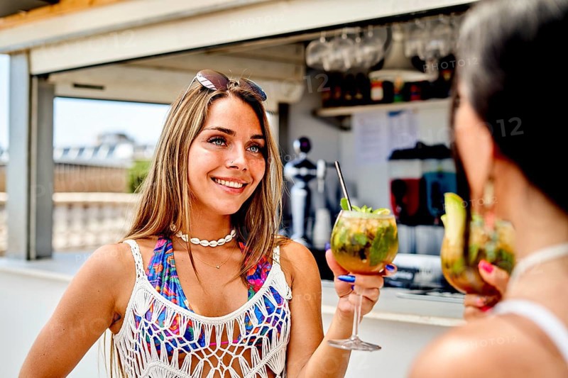 Two friends are at a vibrant beach bar during a sunny summer day, holding colorful cocktails and sharing smiles. The atmosphere is relaxed and filled with joy