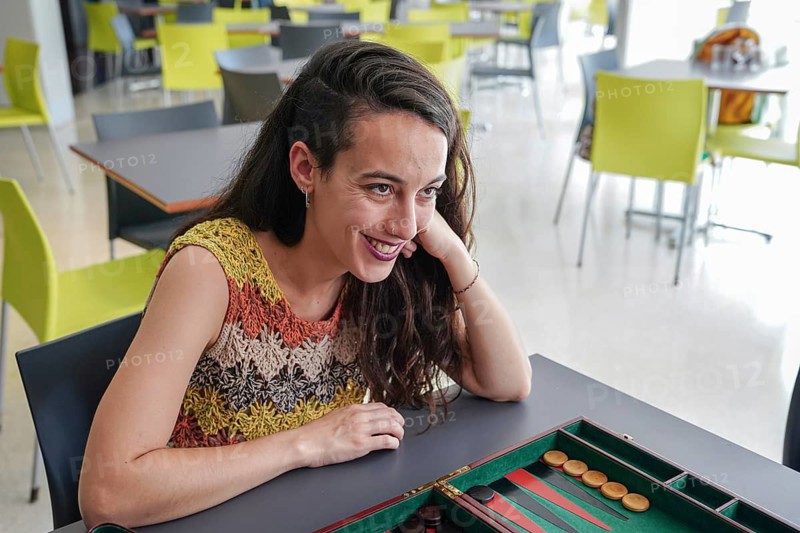 A woman sits at a table in a modern cafe, smiling and engaged, while playing a game of backgammon Bright chairs and natural light create a lively atmosphere