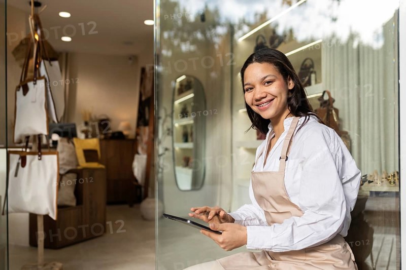 A friendly employee, wearing an apron, sits in a chic bag store, happily using a tablet. The store showcases a variety of elegant bags and a modern interior design
