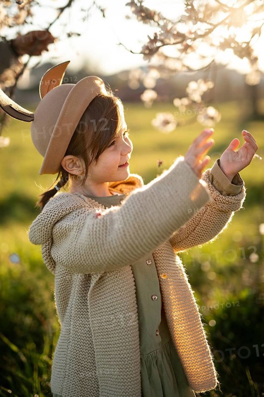 A young girl in a stylish hat smiles as she touches almond blossoms on a sunny spring day