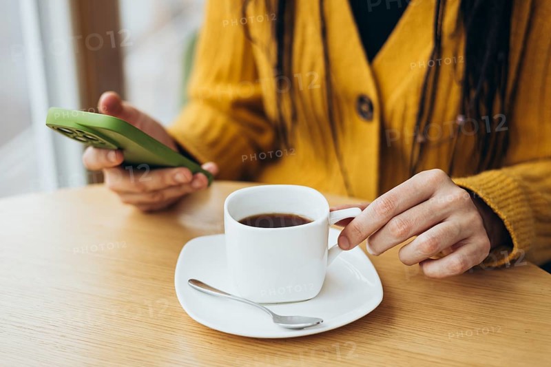 A person in a yellow sweater holds a smartphone while enjoying coffee at a wooden table A white cup, saucer, and spoon create a calming atmosphere