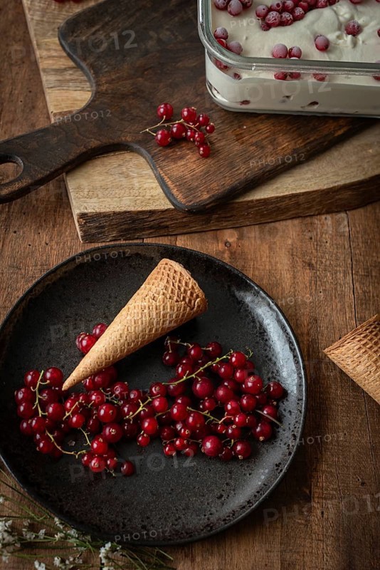 Top view of a rustic food scene featuring red currants on a dark plate beside a waffle cone, set against a backdrop of a wooden board and a glass dish filled with ice cream sprinkled with berries