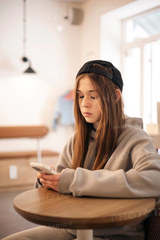 A teenage girl wearing a hoodie and cap sits at a small table, engrossed in her smartphone. The warm, softly-lit environment suggests a cozy, relaxed atmosphere