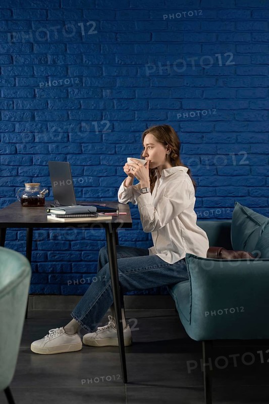 A woman in casual attire sips tea, seated at a table with a laptop, against a vibrant blue brick wall. The modern setting exudes a relaxed yet productive atmosphere