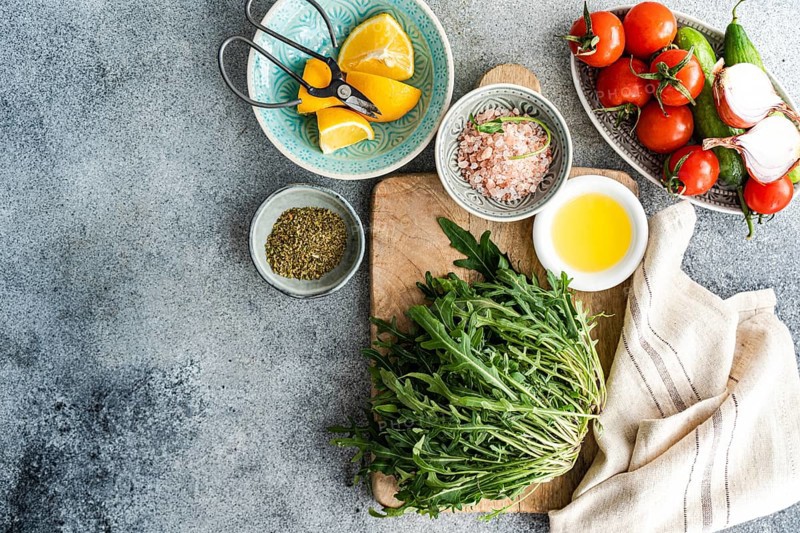Fresh and healthy lunch setup with ingredients including rocket, chopped oregano, cherry tomatoes, cucumber, Himalayan salt, red onion, lemon, and olive oil