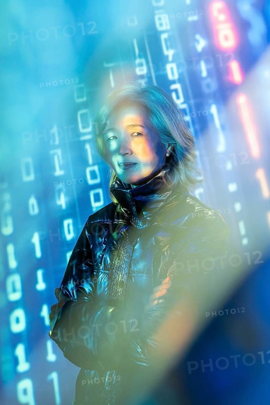An elderly Asian woman stands confidently in the metaverse, surrounded by virtual elements and digital data streams. Her expression reflects curiosity and engagement with technology