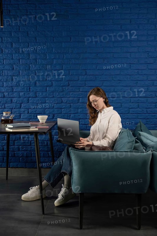 A woman in casual attire works on a laptop, seated on a teal sofa against a blue brick wall. A table with tea and notebooks complements the cozy atmosphere