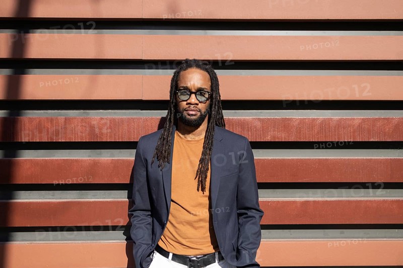 Stylish cuban business man with dreadlocks in sunglasses and casual blazer stands against modern striped wall under sunlight, exuding confidence and urban elegance