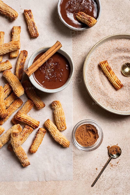 Top view of homemade churros generously dusted with sugar and cinnamon, accompanied by a rich chocolate dipping sauce and a sprinkle of sea salt, all presented on a textured ceramic background for a moody feel