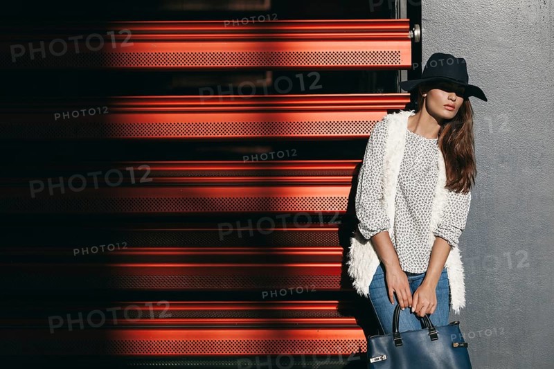 A fashionable woman in a wide-brimmed hat and stylish clothing poses with a handbag against a red background, casting dramatic shadows and exuding urban chic
