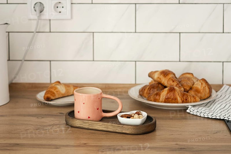 Freshly baked homemade croissants served on a wooden table with a cozy pink mug and a small bowl of nuts. A perfect start to a relaxing morning at home