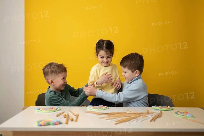 Three children engage in a playful educational lesson in a vibrant yellow classroom. Smiling faces and colorful clay crafts create an atmosphere of joyful learning and imagination
