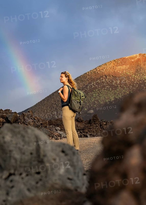 A woman with a backpack explores the volcanic landscape of Timanfaya National Park in Lanzarote. A vivid rainbow arches across the dramatic sky, highlighting the natural beauty
