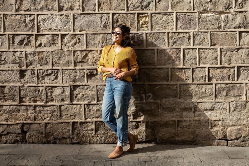 A Latin woman in casual attire, wearing sunglasses, smiles while leaning against a sunlit brick wall. Her relaxed pose and cheerful expression convey a sense of joy