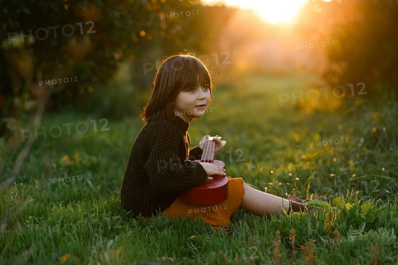 A young child sits on lush grass with orange trees, playing a red ukulele in a sunlit meadow. The warm glow of sunset surrounds the peaceful scene, evoking joy and tranquility