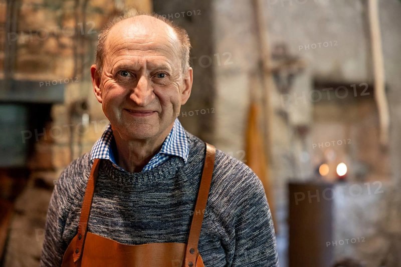 A skilled blacksmith wearing a leather apron smiles warmly in his rustic workshop. The background showcases a traditional forge, tools, and a cozy atmosphere filled with craftsmanship