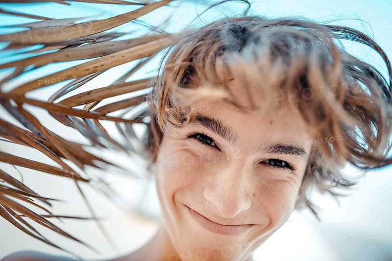 A joyful teenage boy smiles under the dappled shade of a palm tree, his hair tousled by a gentle breeze. The bright blue sky in the background enhances his cheerful demeanor