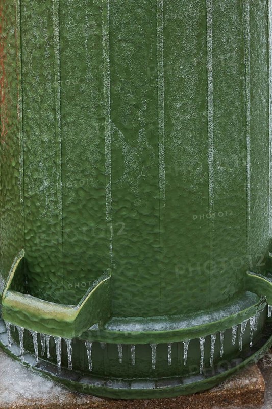 Upside down green PVC and rubber thrash can covered with thick sheet of ice and hanging icicles after freezing rain in late winter, Quebec, Canada
