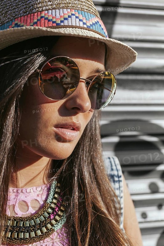 A fashionable woman wearing a straw hat and chic sunglasses. Her long hair and decorative necklace complete the stylish summer look. The background features soft, blurred urban details
