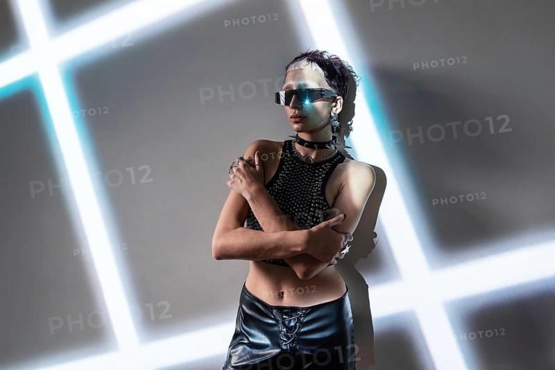 A woman embodies a cyberpunk aesthetic, wearing reflective sunglasses and a studded outfit. The futuristic setting features neon lights, highlighting an edgy, tech-inspired vibe