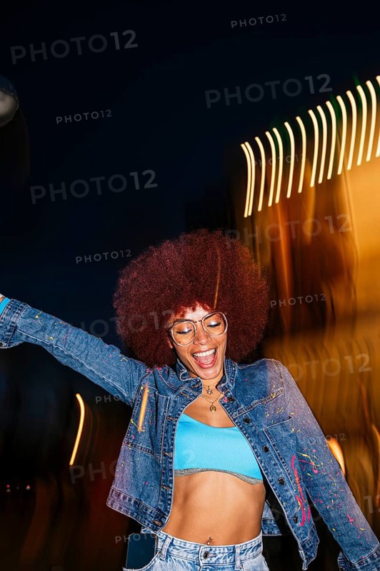 African descendant woman with vibrant afro hair joyfully dancing under night lights. Wearing glasses and a denim jacket, she embodies a lively urban scene with eyes closed