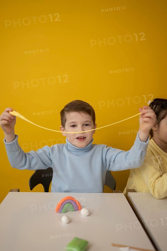 A child joyfully stretches colorful modeling clay during a creative lesson. The bright yellow background enhances the playful atmosphere, promoting imagination and expression