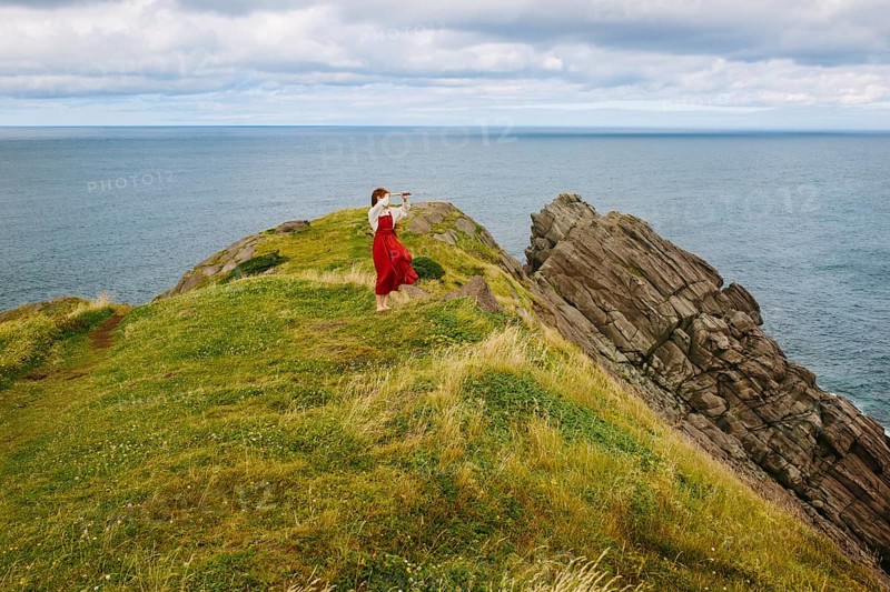 A woman clad in a flowing red dress stands on the rugged cliffs of Middle Cove overlooking the Atlantic Ocean, Newfoundland & Labrador. The image captures a serene moment against a scenic natural backdrop