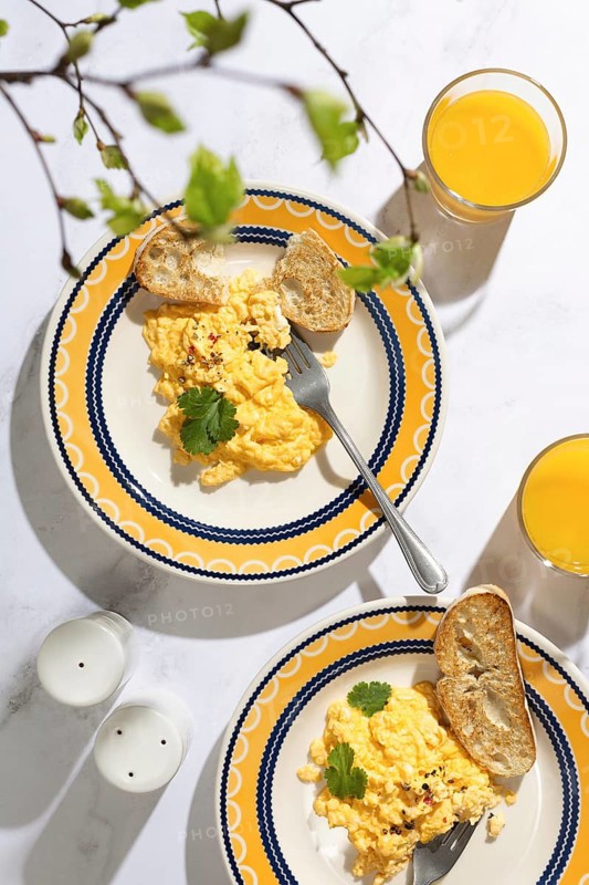 Two plates of creamy scrambled eggs with toast, garnished with herbs, served alongside glasses of fresh orange juice. A perfect, bright morning setup