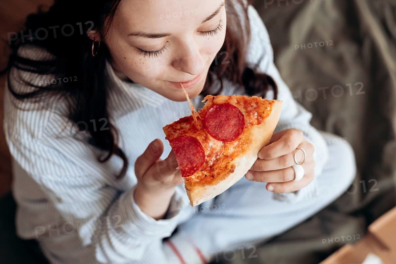 From above image of woman captured in the comfort of her bed savoring a delicious slice of pepperoni pizza, highlighting the joys of casual indoor dining