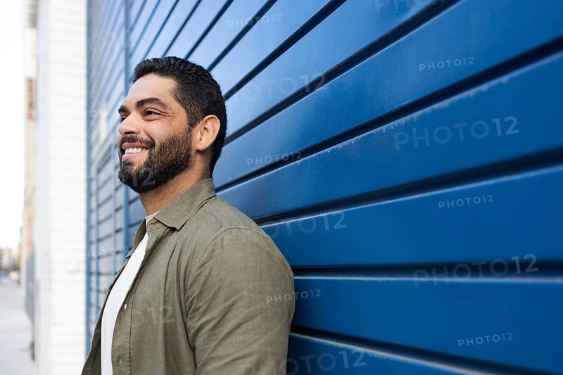 A bearded man with a friendly smile leans against a blue wall on an urban street. He has short hair and wears a casual green shirt over a white t shirt, exuding confidence in the city