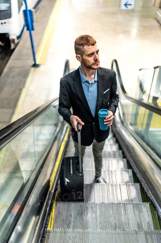 Businessman going up escalator in subway station, holding reusable coffee cup and pulling trolley suitcase