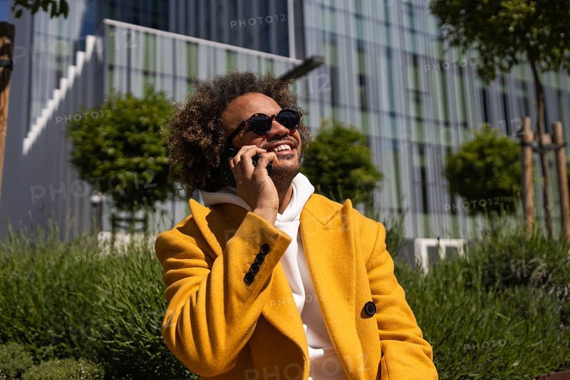 Afro man wearing a vibrant yellow coat, smiling while talking on his phone outdoors. He is seated amidst greenery, with a modern building in the background