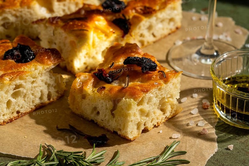 Golden homemade focaccia topped with sun dried tomato and rosemary. Served with olive oil, this savory delight rests on parchment paper, offering a rustic and appetizing look