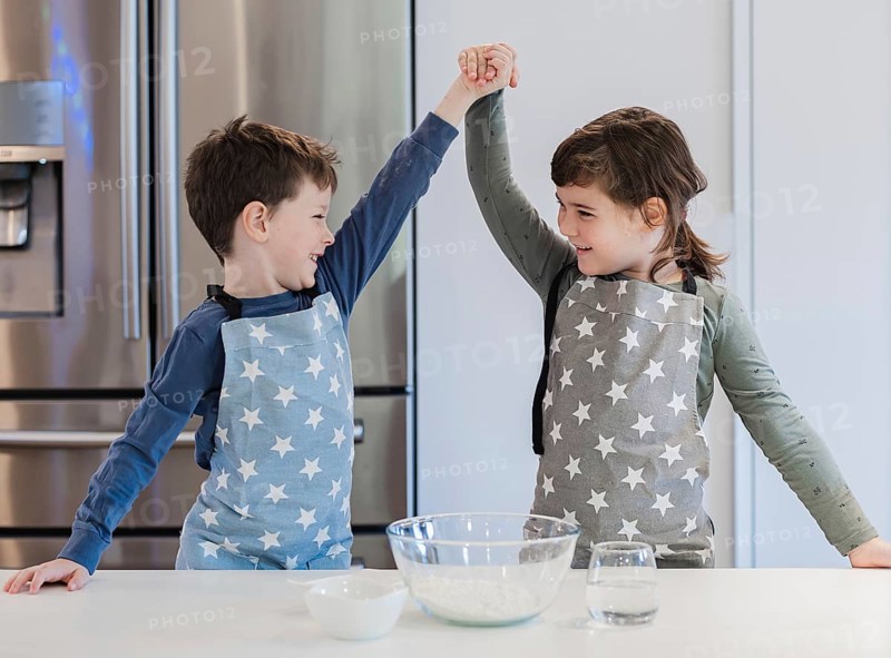 Two young siblings in star patterned aprons enthusiastically raise their hands in celebration while baking in a modern kitchen, capturing joyful teamwork and family bonding