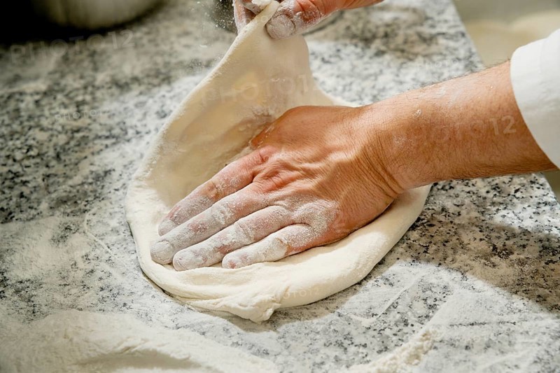 Cropped unrecognizable person kneading fresh pizza dough on a flour-dusted marble countertop, focusing on handwork and ingredients in the kitchen