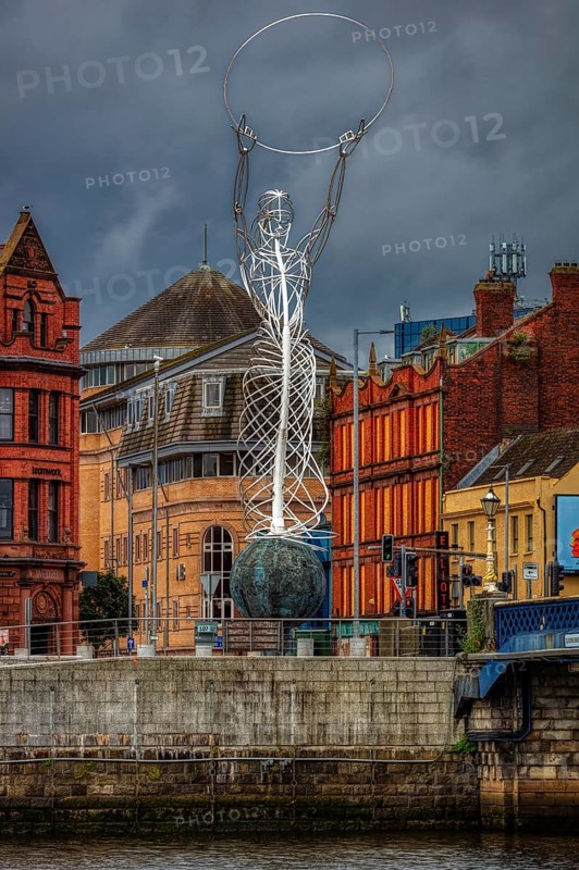 Modern metal sculpture on a riverbank in front of historic buildings under a cloudy sky, Belfast