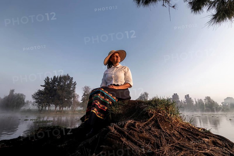 A woman in traditional attire enjoys a peaceful moment by the water in Xochimilco, Mexico. Surrounded by nature, she reflects the region's rich cultural heritage and tranquil beauty