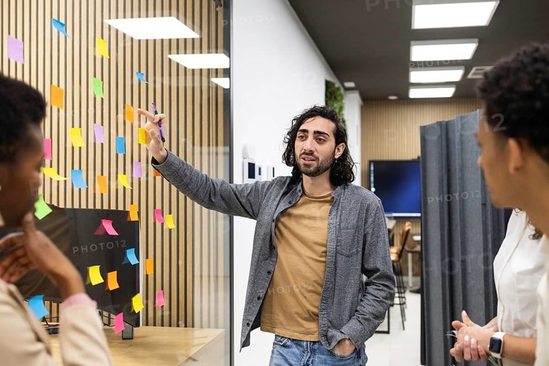 Professionals engage in a brainstorming session in a modern office space. A man gestures towards colorful sticky notes on glass, fostering creativity and collaboration