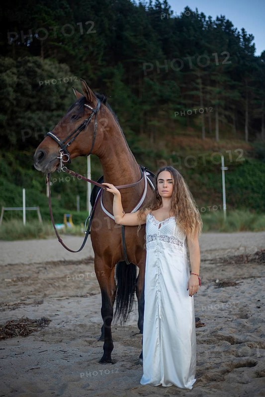 A young woman in a flowing white dress stands beside her horse on a tranquil beach. The scene captures a blend of elegance and natural beauty, with a serene forest backdrop