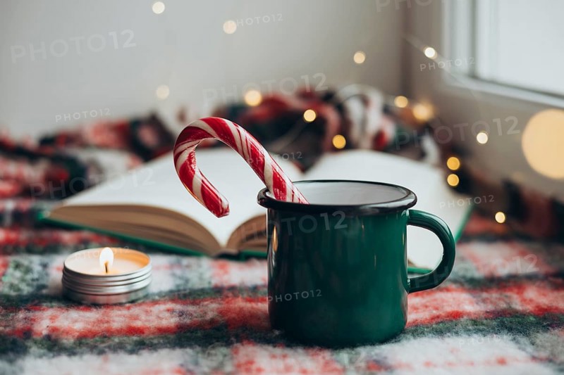 Cozy Christmas setting featuring a green mug, a festive candy cane, and a flickering candle next to an open book on a plaid blanket, creating a warm holiday atmosphere