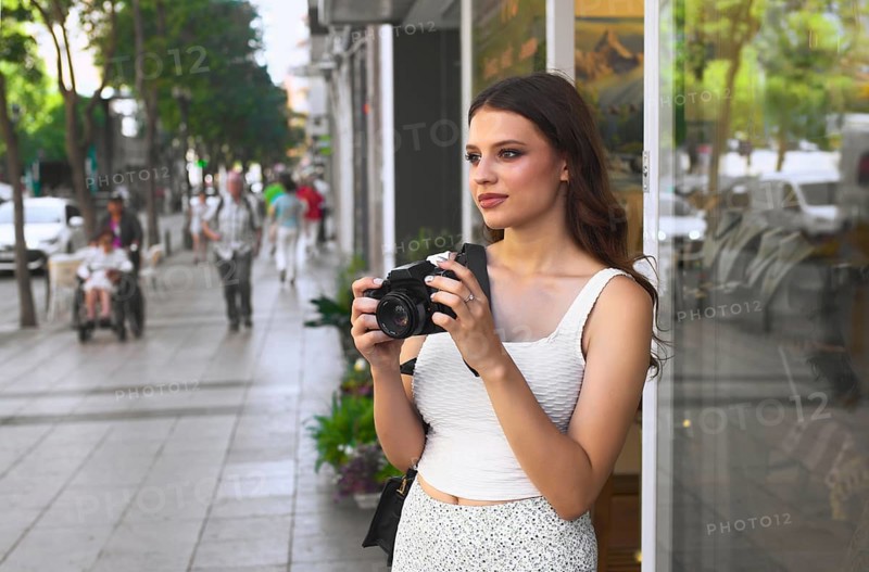 A young woman stands on a bustling city street, holding a camera She gazes ahead, ready to capture moments amidst the vibrant urban scene Pedestrians walk past her