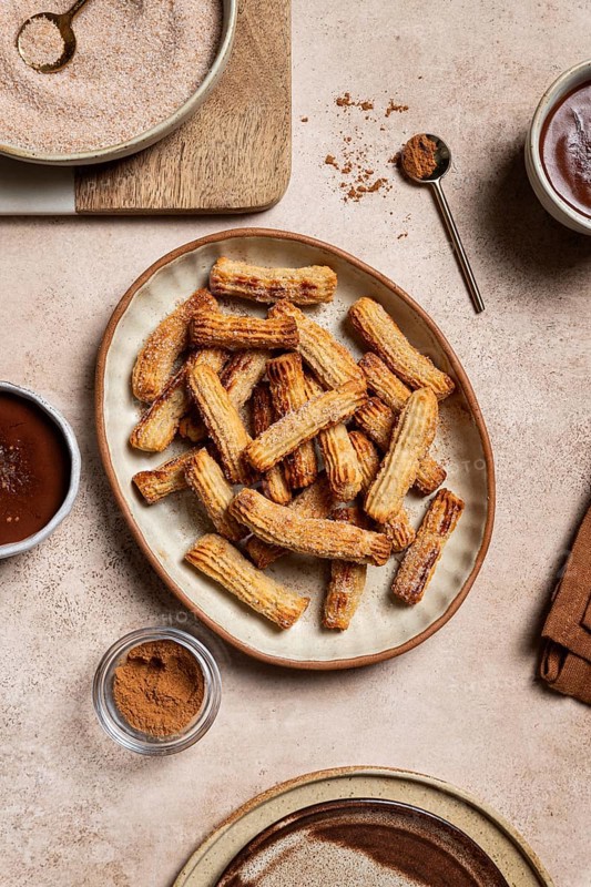 Top view of a ceramic plate filled with sugary churros next to bowls of chocolate sauce and cinnamon, set against a background of rustic linen and wooden kitchenware